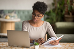 Woman with glasses looking at laptop while writing in notebook